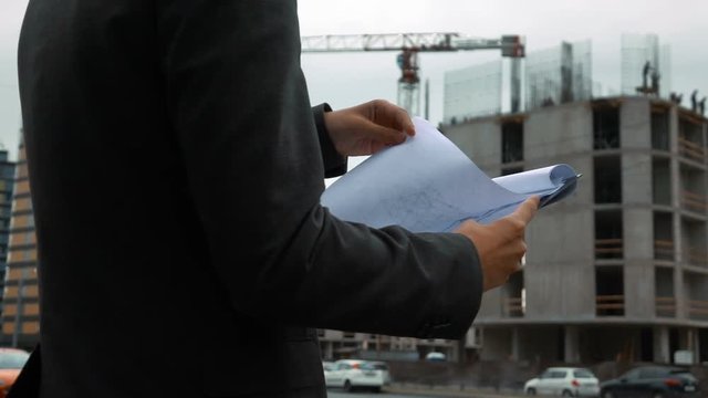 Slow motion. Project manager checking clipboard construction plan with newly constructed building with crane and beams at background. Detailed close up of text pad.
