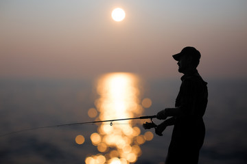 fisherman at dusk. silhouette of a fisherman with a fishing rod in the background reflected in...