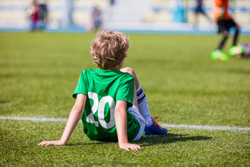 Fototapeta premium Youth Soccer; Little Soccer Player. Young Boy as a Soccer Player Sitting on Football Field and Watching School Soccer Match