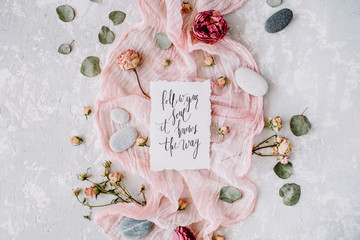 inspirational quote "follow your soul it knows the way" written in calligraphy style on paper with dry white tulips, eucalyptus petals and pink textile on concrete background. Flat lay, top view