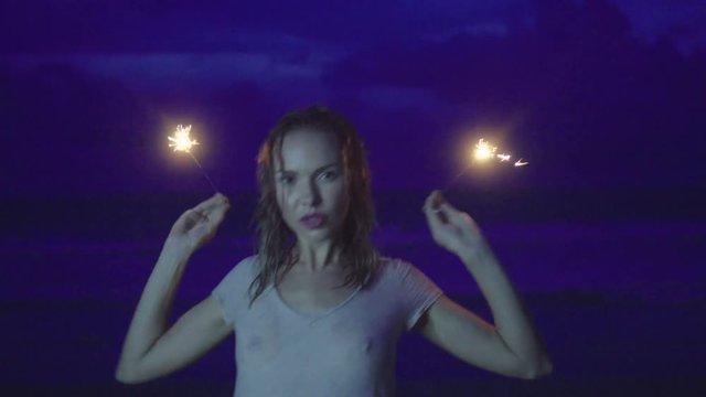 Closeup of sensual woman wearing t-shirt with wet hair doing sparkler fireworks show on the beach at night over sea and sky background