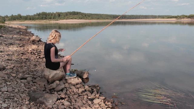 Girl Catches Fish. Girl sitting on a rock by the river. Girl holding a fishing rod. Girl catches fish in the river
