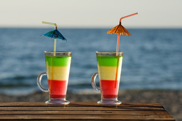 A pair of colorful cocktails on a wooden table on the beach, the sea in the background