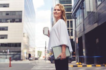 Beautiful young businesswoman with coffee holding laptop on city background