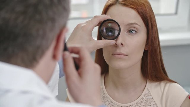 Closeup tilt up of optometrist checking eyes of female patient through lens