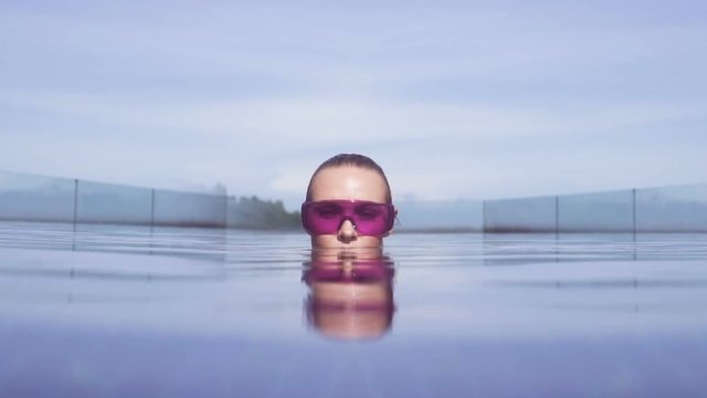 Face closeup of sexy young woman wearing purple sunglasses looking at camera in infinity rooftop swimming pool on a sunny day over blue sky