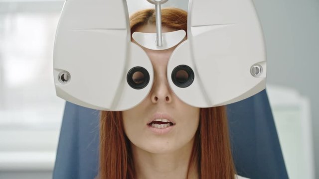Closeup of young red-haired woman reading eye chart during distance vision exam with modern automated phoropter