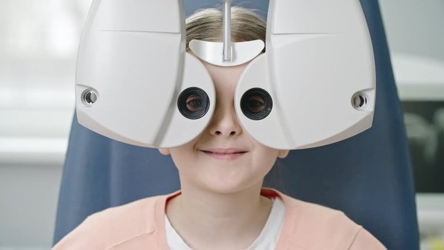 Closeup of little girl reading eye chart during distance vision exam with modern automated phoropter and then smiling at camera