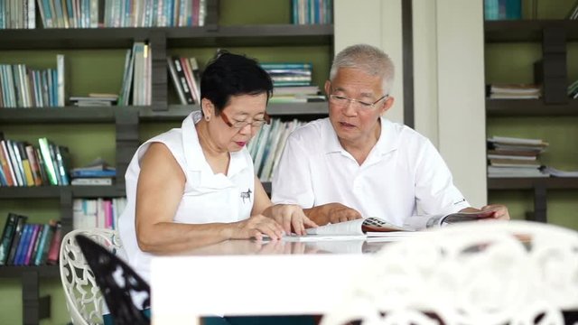 Asian senior couple reading books and magazines together in small library. Learning and study concept
