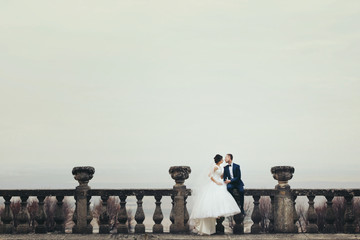 Bride and groom sit on the stone balcony with a deep colored sky