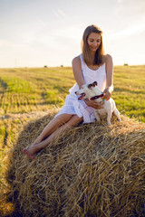 Naklejka na ściany i meble young girl in a white dress is sitting with Jack Russell Terrier dog on the haystack