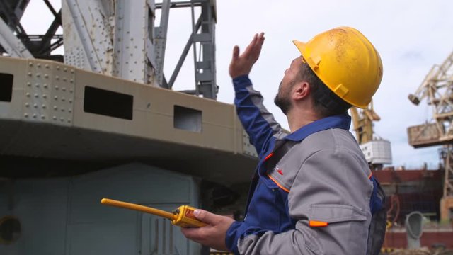 Dock worker with yellow helmet and walkie-talkie directs the cargo crane in Shipping port
