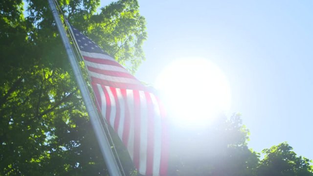 American Flag Slow Sun Flare. an american flag waves in front of the sun flare with a tree in the background
