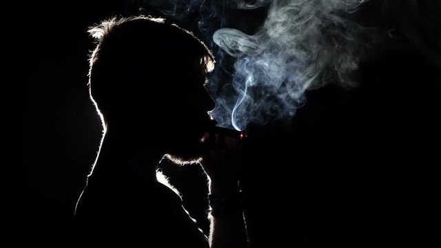 Brutal man smokes a cigar. Takes a big drag and puffs smoke. Blacklight. Silhouette. Close up. Slow motion