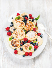 Syrniki or cottage cheese pancakes with fresh forest berries and sour cream sauce in serving dish over white wooden background, selective focus. Russian and Ukranian cuisine