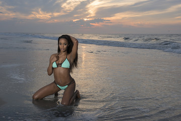 Fototapeta na wymiar Beautiful African American female model posing on beach in swimsuit at sunrise (sun rising behind her over ocean) - turquoise colored bikini