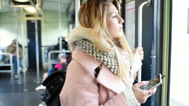 Half length of young handsome blonde straight hair woman traveling in the underground, holding a smart phone, looking outside the window - technology, commuting, transport concept