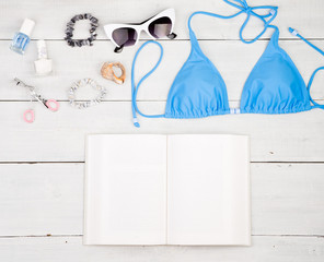 blue swimsuit, book, sunglasses, cosmetics makeup, bijou and essentials on white wooden desk