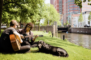 Two young men playing acoustic guitar in a city park in the company of a black dog. Selective focus.