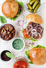Homemade beef burgers with onion, pickles, fresh vegetables, spices, sun-dried tomatoes and tomato sauce on serving wooden board over white painted background. Top view, vertical composition