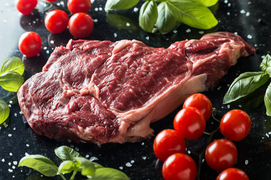 Fresh steak served with spices, tomatoes and leafs of basil on marble background. Uncooked beefsteak cooking on a kitchen. Delicious, spicy, juicy meat with copy space closeup.
