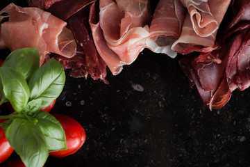 Jambon mix. Ham. Traditional Italian and Spanish salting, smoking, dry-cured dish - jamon Serrano and prosciutto crudo sliced with herbs and tomatoes on dark stone background. Copy space. Closeup. 
