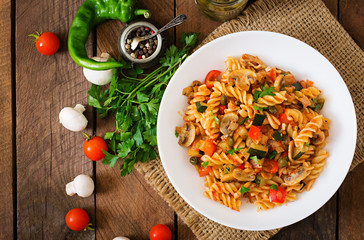 Vegetarian Vegetable pasta Fusilli with zucchini, mushrooms and capers in white bowl on wooden table. Top view