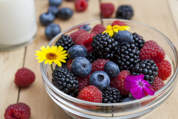 Glass bowl of fresh blackberries, raspberries, blueberries and marigold on light wooden table