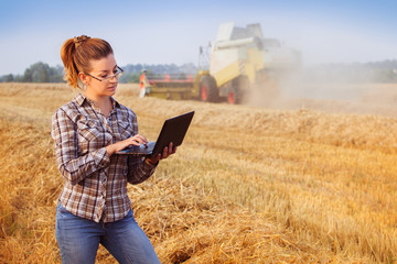 Young pretty farmer girl in glasses with hair tied in a ponytail make notes on notebook in wheat field while combine harvester working in background. Image released.
