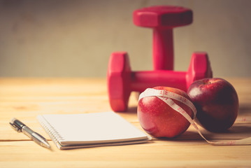 Apple, tape measure and notepad with dumbbells lying on a wooden