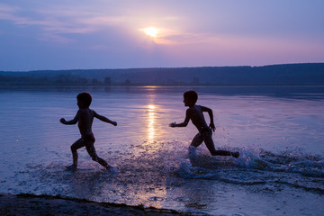 Fototapeta premium Two boys running on river's beach against sunset