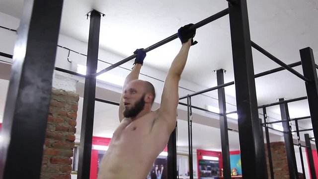 Shirtless muscular bearded male doing exercises on horizontal bar in a gym club.