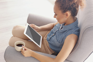 Beautiful African American girl with coffee and tablet on armchair