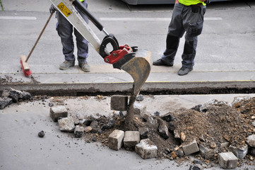 Straßenbau - Baustelle: Bagger gräbt Grube und hebt Pflastersteine aus dem Asphalt