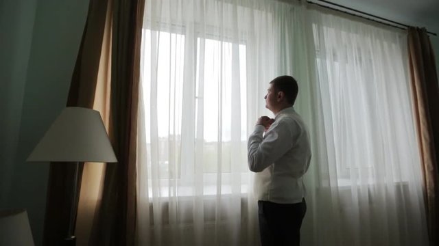 Man in white shirt tying a tie near the window