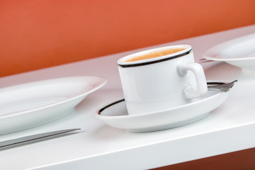 Espresso, Besteck und Teller auf weißem Tisch vor orangenem Hintergrund, Makro