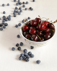 Cherry in a bowl on a table, lies bilberry nearby. A cloth from flax