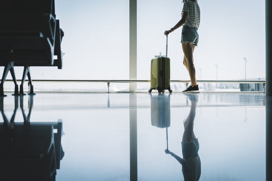 Young hipster girl tourist waiting her flight or boarding at the airport, attractive female traveler standing near a window while waiting for boarding an aircraft, summer vacation concept