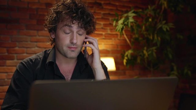 Young successful businessman speaking on phone, smiling, working in office at night. Slow motion.
