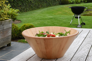 Fototapeta na wymiar Fresh mixed summer salad in wooden bowl. Barbecue unit on green grass in the background.