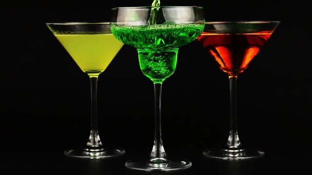 Pouring green cocktail in wineglass. Three glasses of different cocktails edge on black background