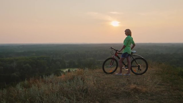 Crane shot: Woman standing near a bicycle looks at sunset