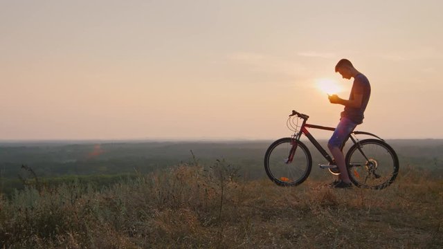 Teenager sitting on the bike uses a smartphone. In the sunset of his silhouette