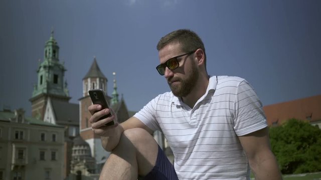 Young man browsing smarthpone, outdoor. He is sitting. He is young, good looking and has beard. Man is dressed in blue shorts, polo shirt and sunglasses.
