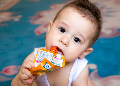 Happy Baby Child Sitting In Chair And Eats Food From A Tube By Yourself, The Kid Was Holding Pack Of Fruit Puree, Lose-up, In The Kitchen, Start Feeding