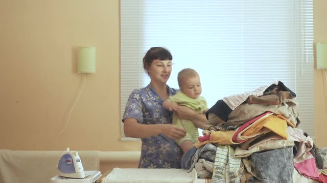 Mother holding baby in arm, ironing with the other arm. In a room near the window.