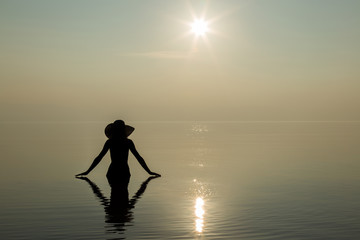 Fototapeta na wymiar Silhouette. Young woman with hat on head enjoy the sunset on the beach and she feels the harmony in herself. Serene and warm, gentle water. Touch to water. In front of view opens infinity.