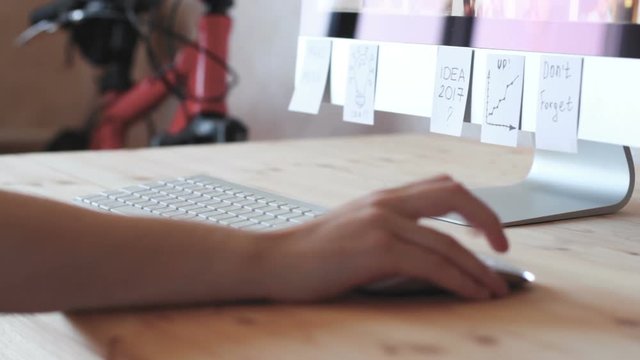 A freelancer working at a computer plastered with notes, reminders, stickers. Scrolling page with mouse closeup.