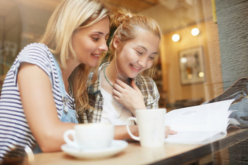 Two young blond girls reading fashion magazine in comfy coffee shop near the window. Nice woman with long hair smiling and the other girl with bun in checked shirt looking at page with pleasure.