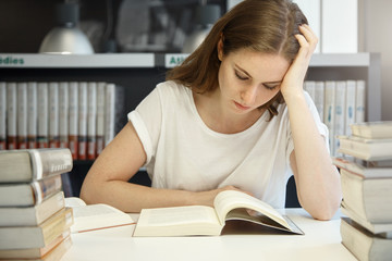 Portrait of pretty university girl reading a textbook on economics with focused and concentrated expression, sitting at the table surrounded with piles of books while preparing for final exams